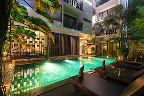 A pool area with lounge chairs and a dining area, surrounded by greenery and a multi-level building.