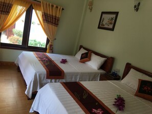Quarto de hotel com duas camas, cada uma com colcha branca e borda decorativa marrom. Há uma janela com cortinas, um quadro na parede e uma pequena mesa com garrafa e copos.