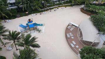 A resort with a sandy beach, palm trees, a pool area, and a deck with lounge chairs.