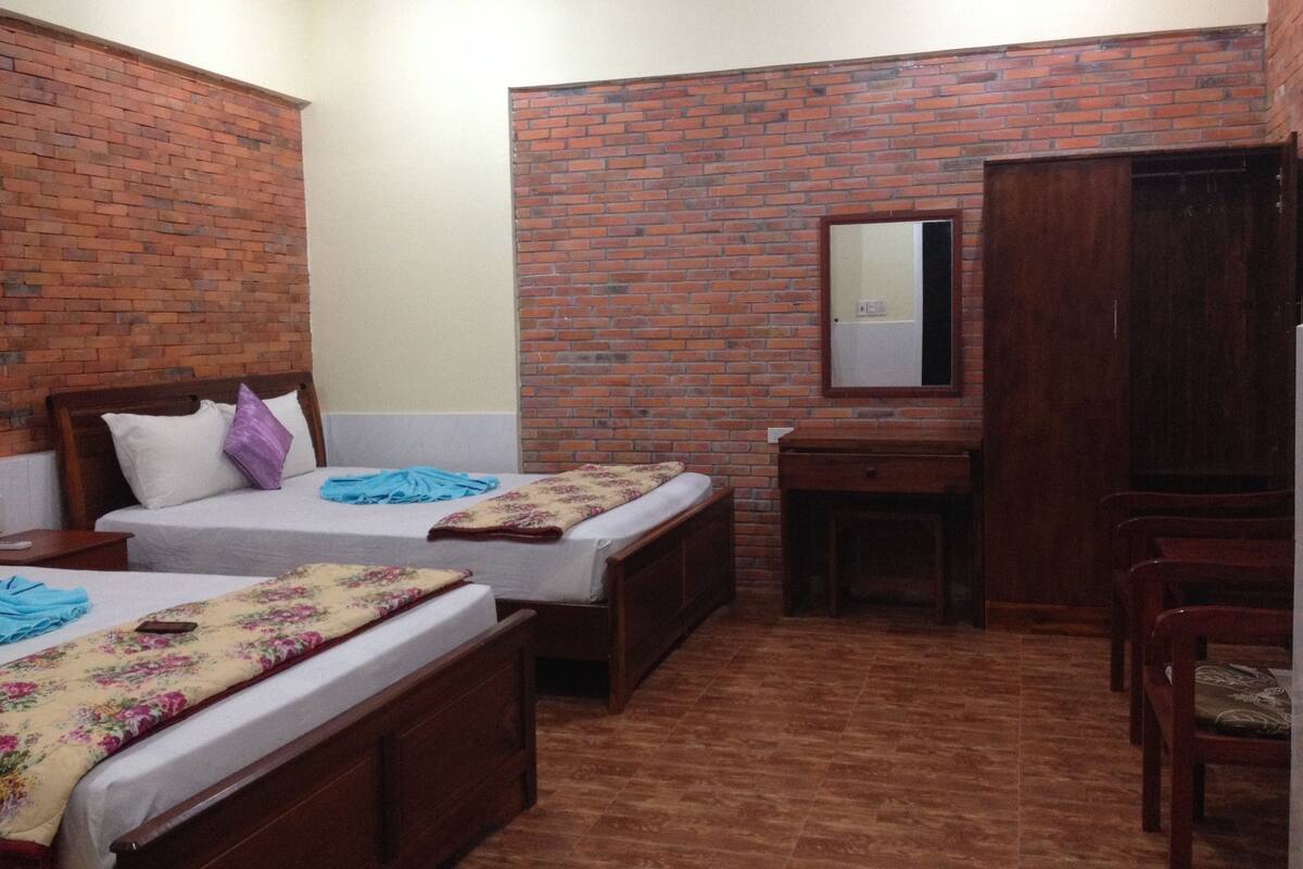 Quarto com duas camas de madeira, uma mesa pequena de madeira, um espelho e uma parede de tijolos.