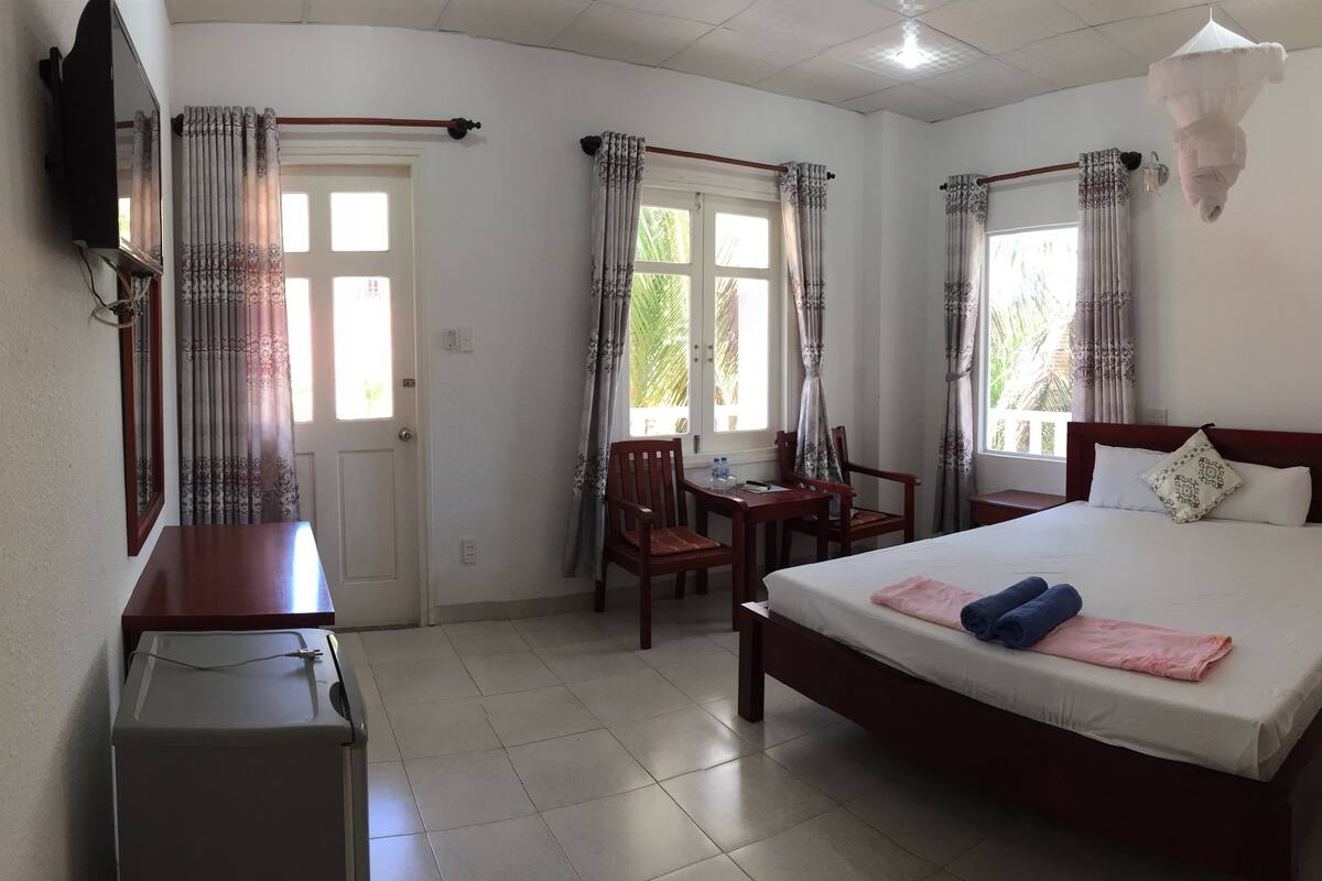 Quarto de hotel com cama, escrivaninha, cadeira, televisão e cortinas.