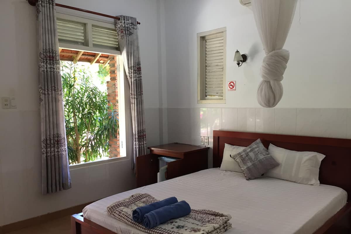 Cama bem arrumada com manta xadrez e toalhas dobradas, mesa de cabeceira, abajur na parede e janela com cortinas.