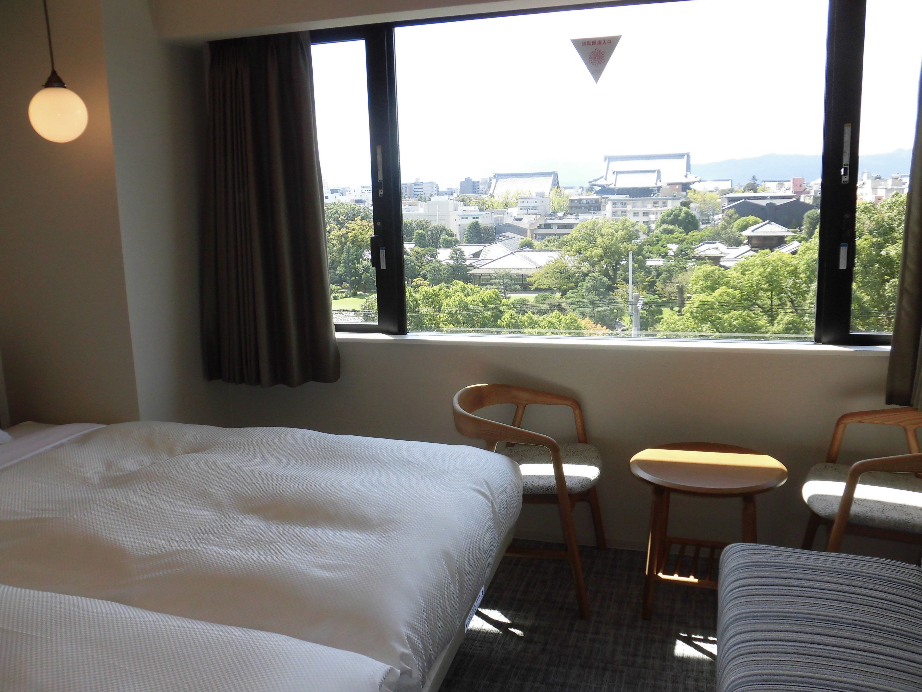 A hotel room with a bed, two chairs, a small table, and a view of a cityscape through large windows.