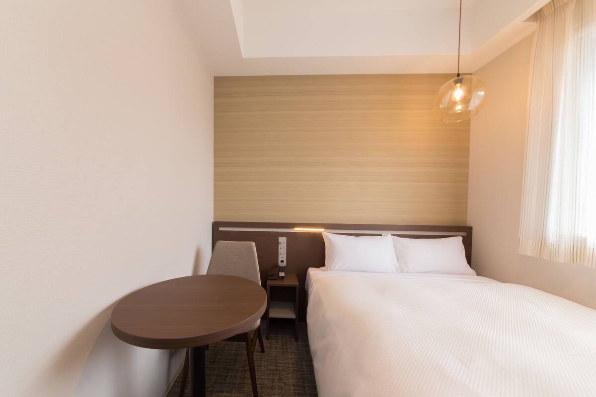 A hotel room with a bed, a small table, a chair, a nightstand, and a wall-mounted light fixture.