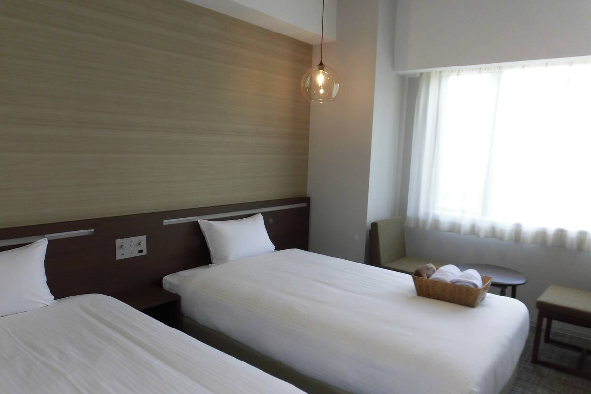 A hotel room with two beds, a small table, a chair, and a window with curtains.