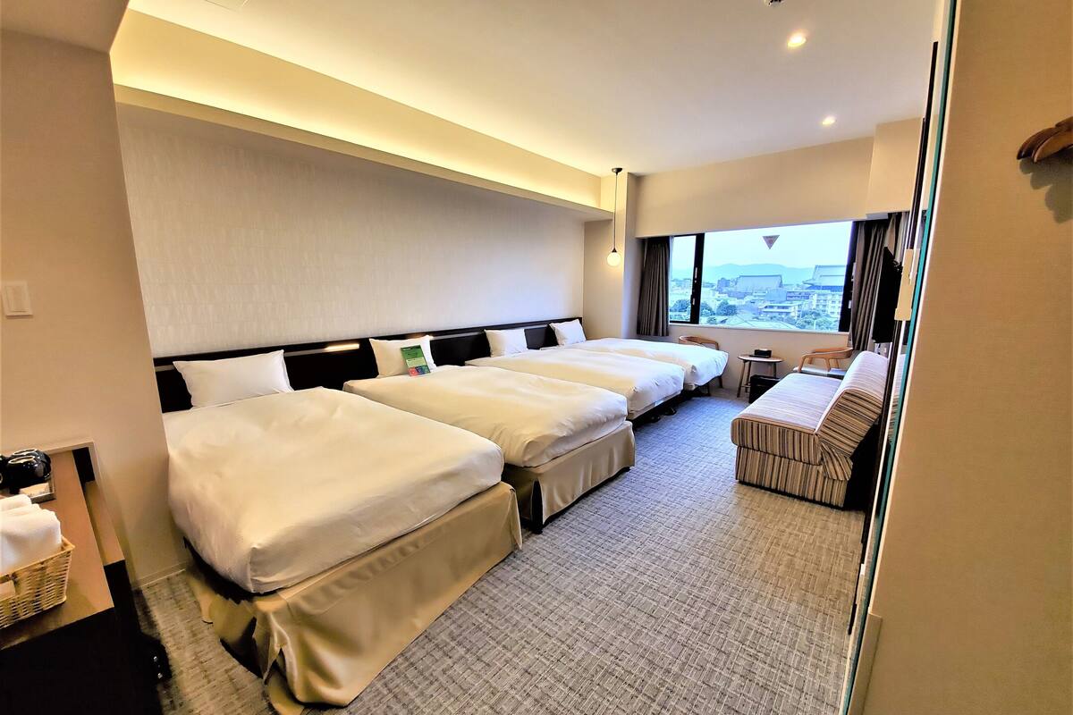A hotel room with three beds, a small table, and a view of the city.