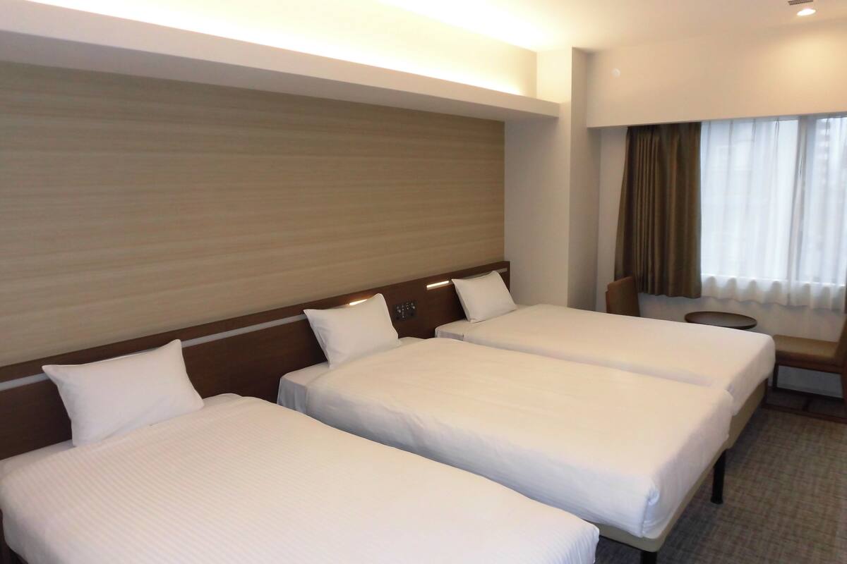 A hotel room with two single beds, a wooden headboard, a small round table, and a window with curtains.