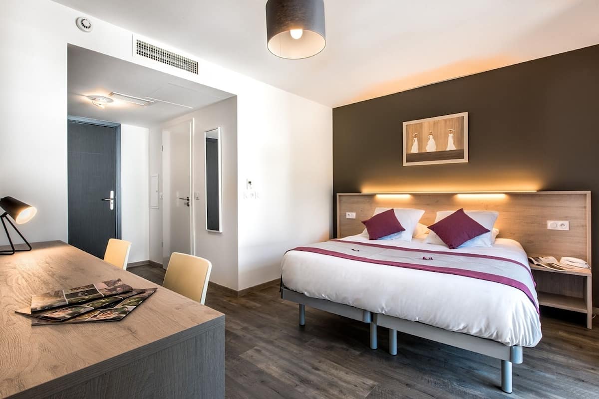 A modern hotel room with a bed, bedside table, and a desk with magazines.