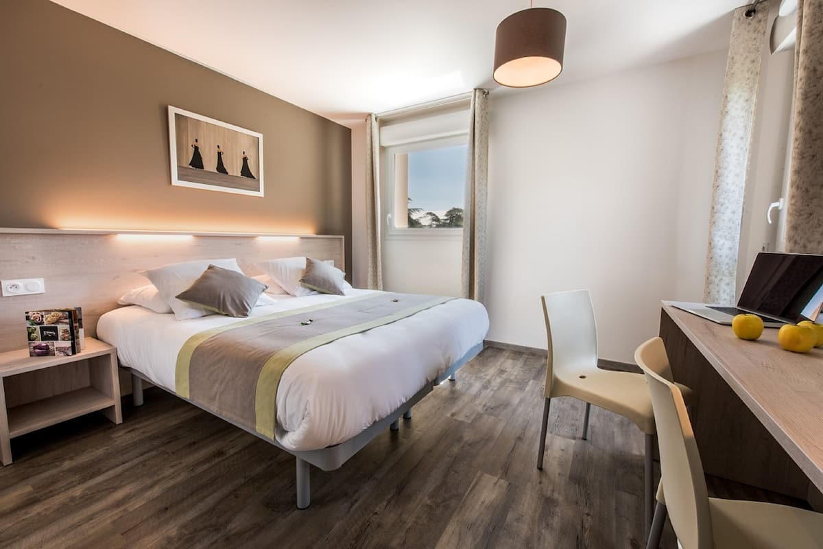 A bedroom with a bed, bedside table, lamp, and a desk with a chair.