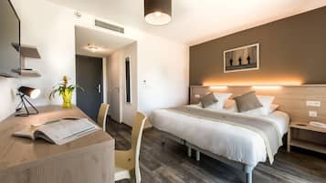 A modern hotel room with a wooden desk, a bed with a headboard, a bedside table, and a wall-mounted artwork.