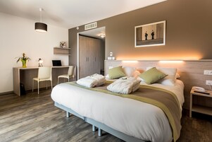 Una habitación de hotel moderna con una cama grande, mesitas de noche, un escritorio con una silla, una planta y un cuadro en la pared.