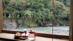 A view of a river with a rocky riverbank and dense greenery. A wooden table with a teapot, cup, and plate of pastries is visible. - 51 Spring (New Taipei City)