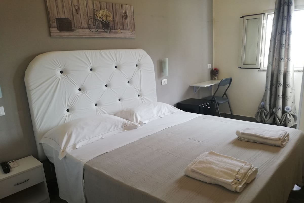 A neatly made bed with a white headboard and pillows, a bedside table with a remote control, and a small desk with a chair.