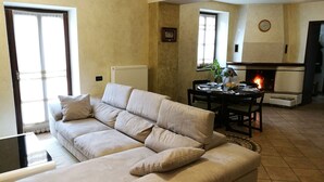A living room with a beige sofa, a dining table set for four, a fireplace, and a large window. - Le Cicogne (Rovereto)