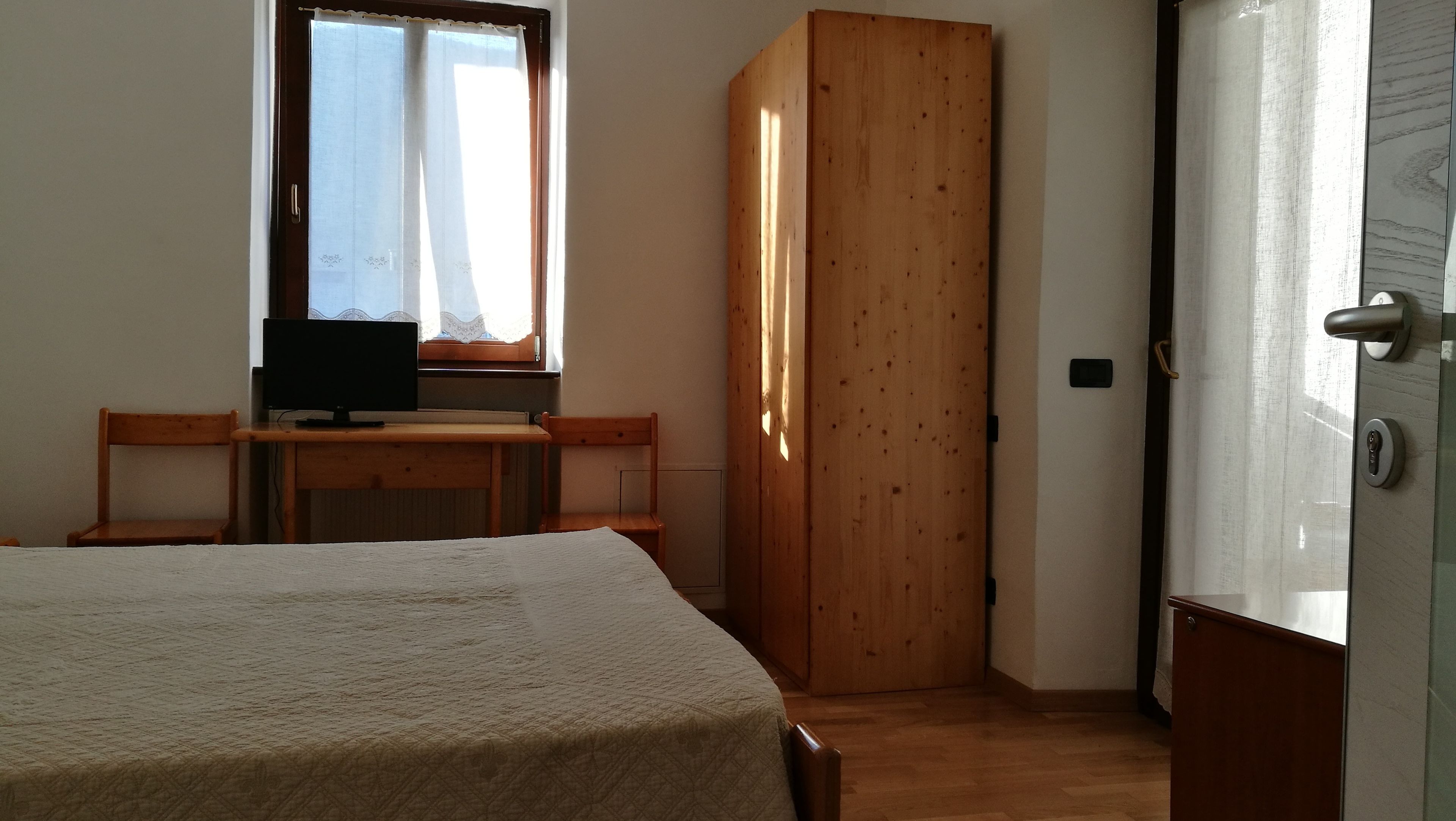 Een slaapkamer met een bed, een bureau met een computer, een raam met gordijnen en een houten garderobekast.