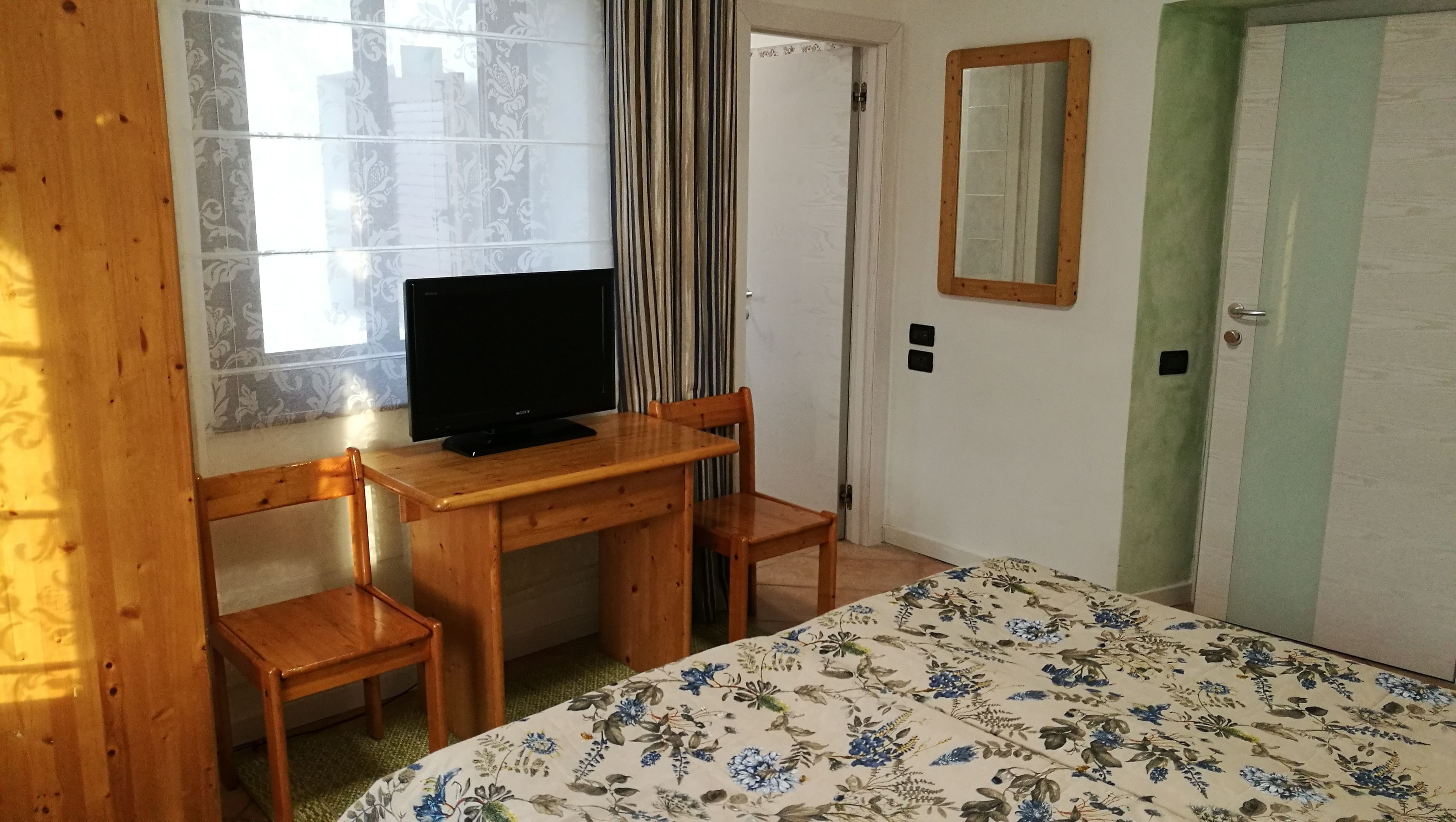 Een kamer met een bed, een bureau met een televisie, twee stoelen, een spiegel en een deur.