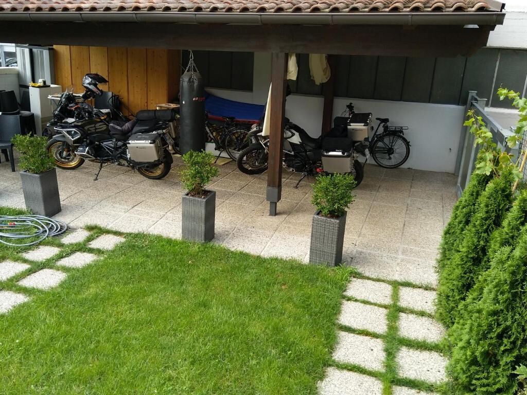 A covered outdoor area with multiple motorcycles parked under a shelter, surrounded by potted plants and a grassy area.