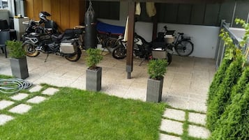 A covered outdoor area with multiple motorcycles parked under a shelter, surrounded by potted plants and a grassy area.