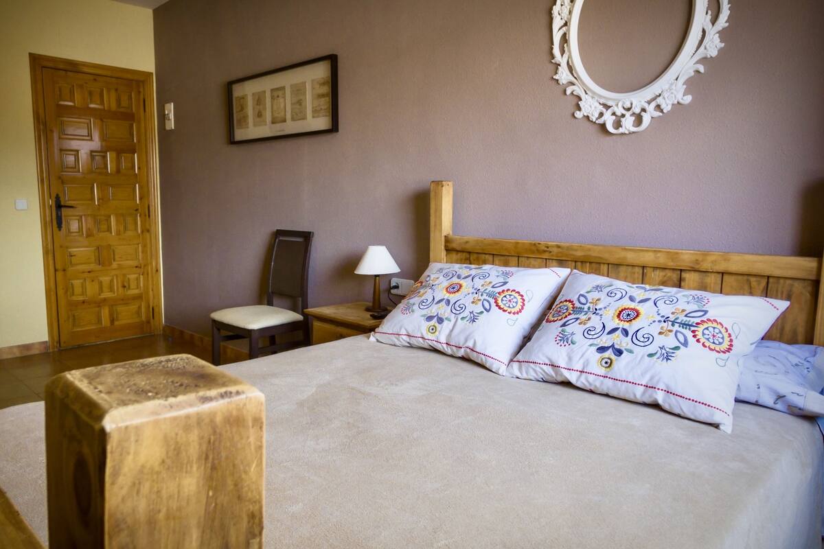 A bedroom with a wooden bed, two pillows with floral patterns, a nightstand, a chair, a wooden bench, a wooden door, and a framed picture on the wall.