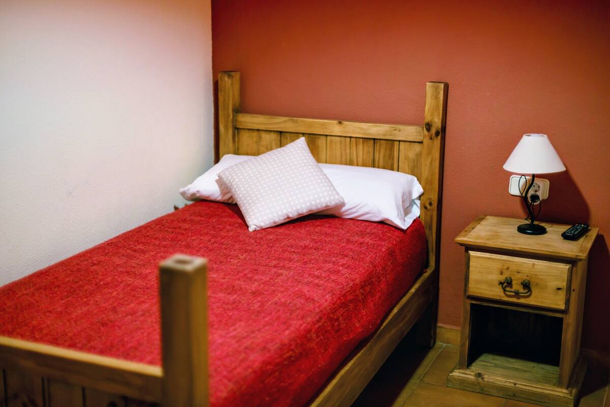A single bed with a wooden headboard, a red bedspread, and a white pillow. There is a small wooden nightstand with a lamp and a remote control.