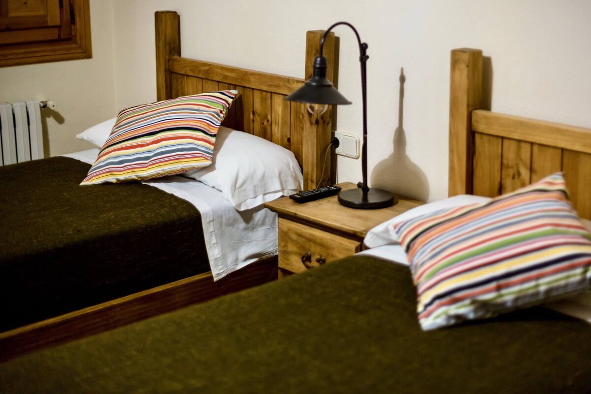 A room with two single beds, each with a striped pillow and a white pillow. There is a wooden nightstand between the beds with a lamp and a remote control.