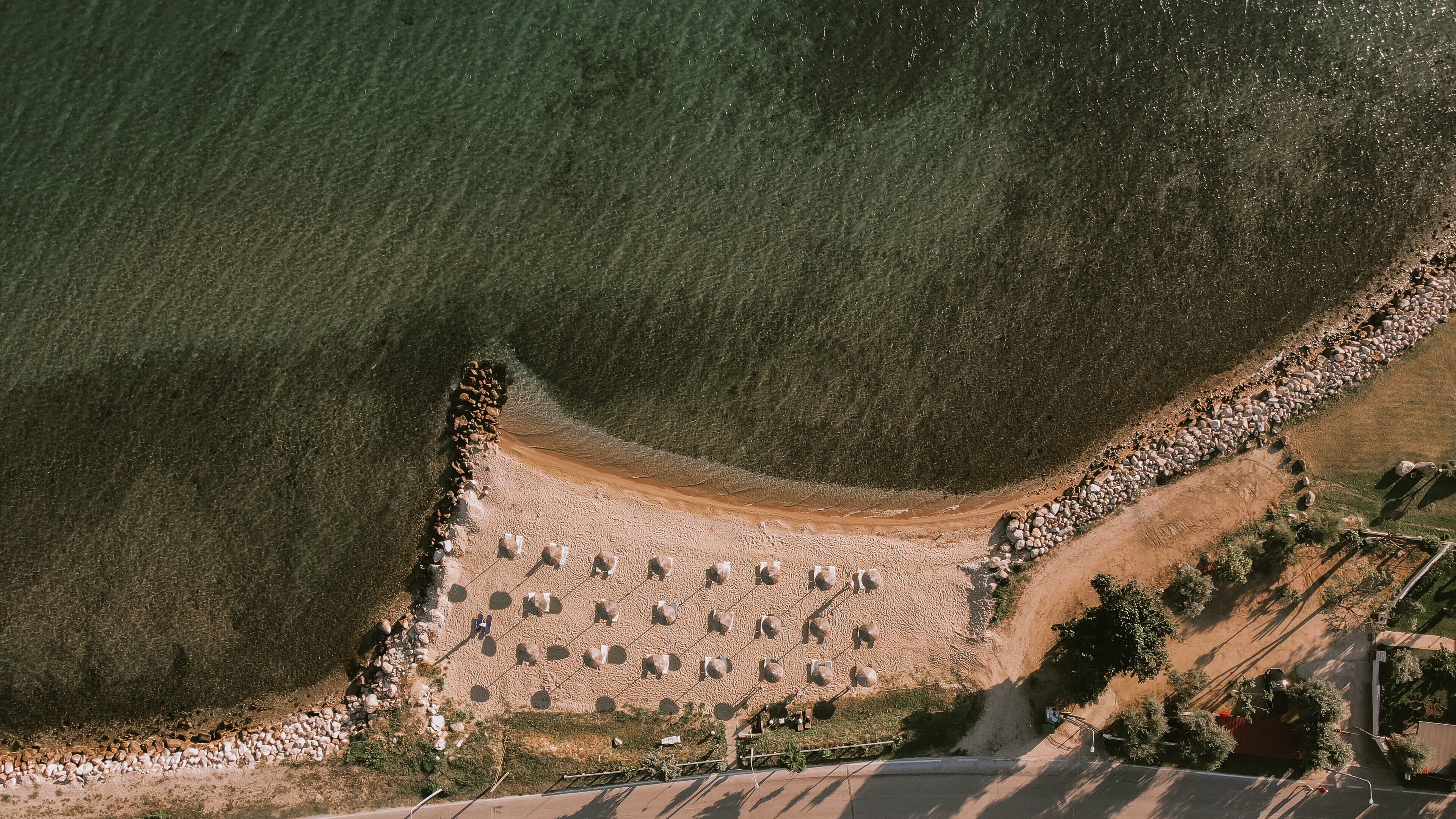 Eine Vogelperspektive einer Küstenlandschaft mit Sandstrand, Steinmauer und einer Straße, die zu einem Gebäude führt.