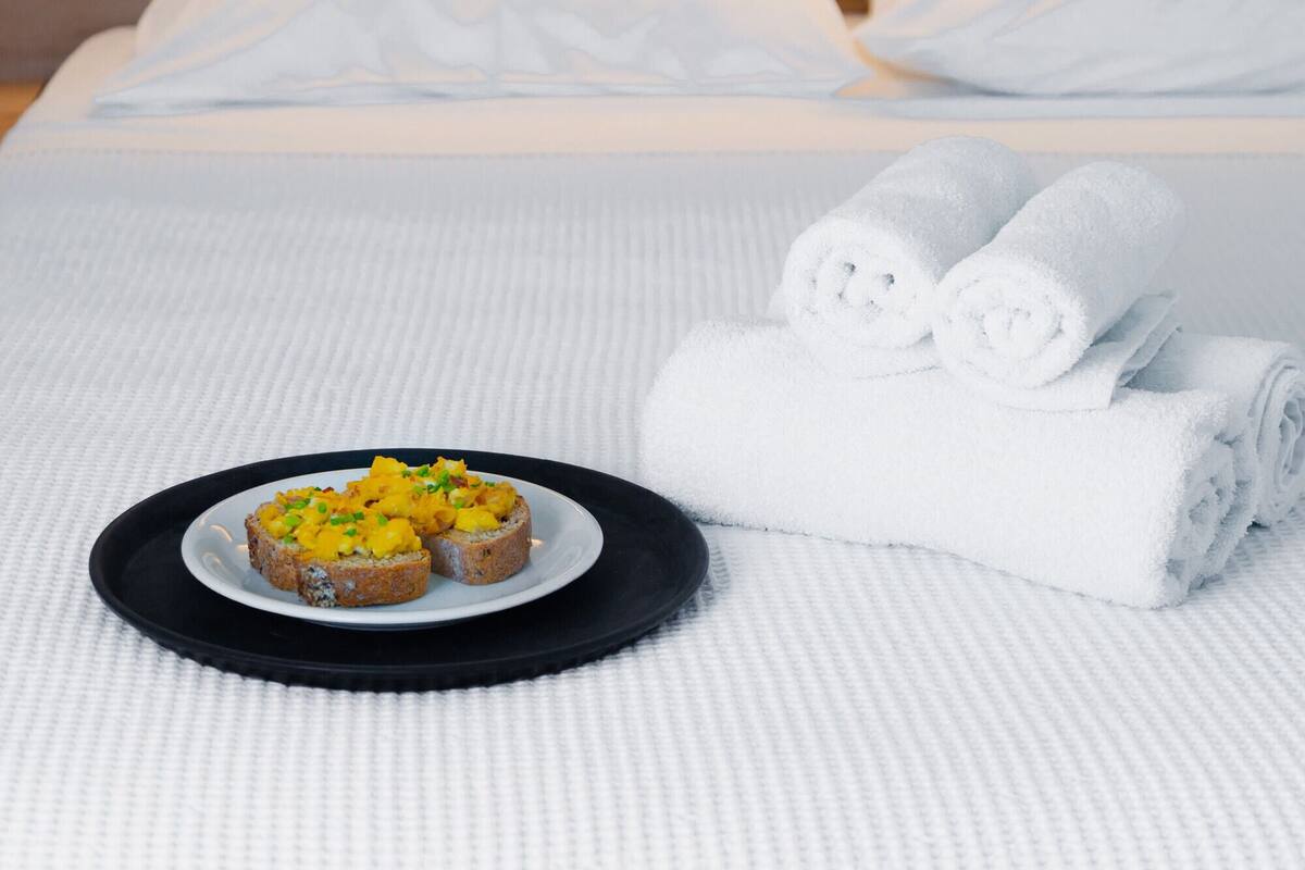 Ein ordentlich gemachtes Bett mit weißen Leinen, ein Teller mit Speisen und aufgerollte Handtücher.