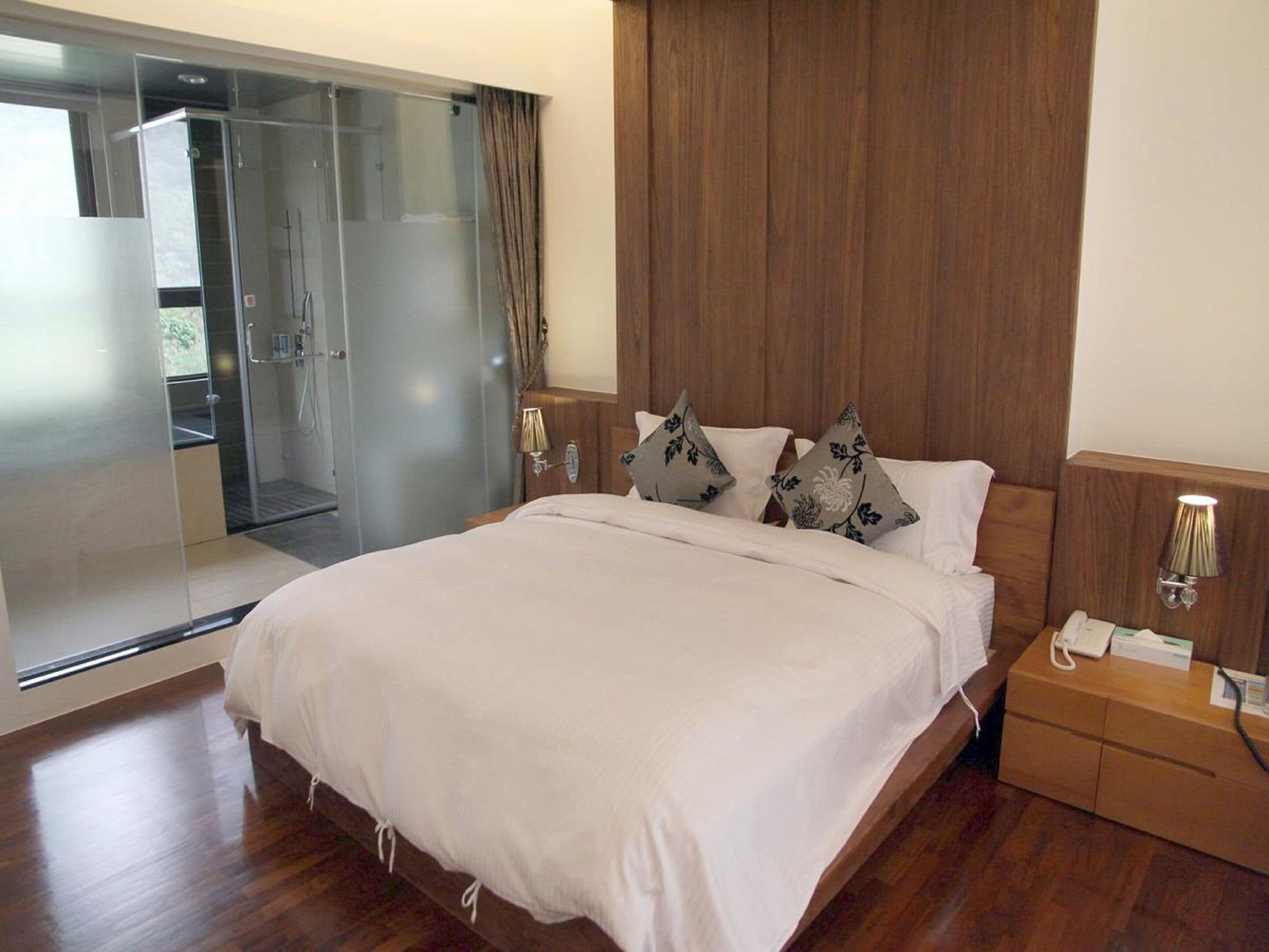 A modern bedroom with a large bed, a wooden headboard, a glass shower enclosure, and a bedside table with a lamp.