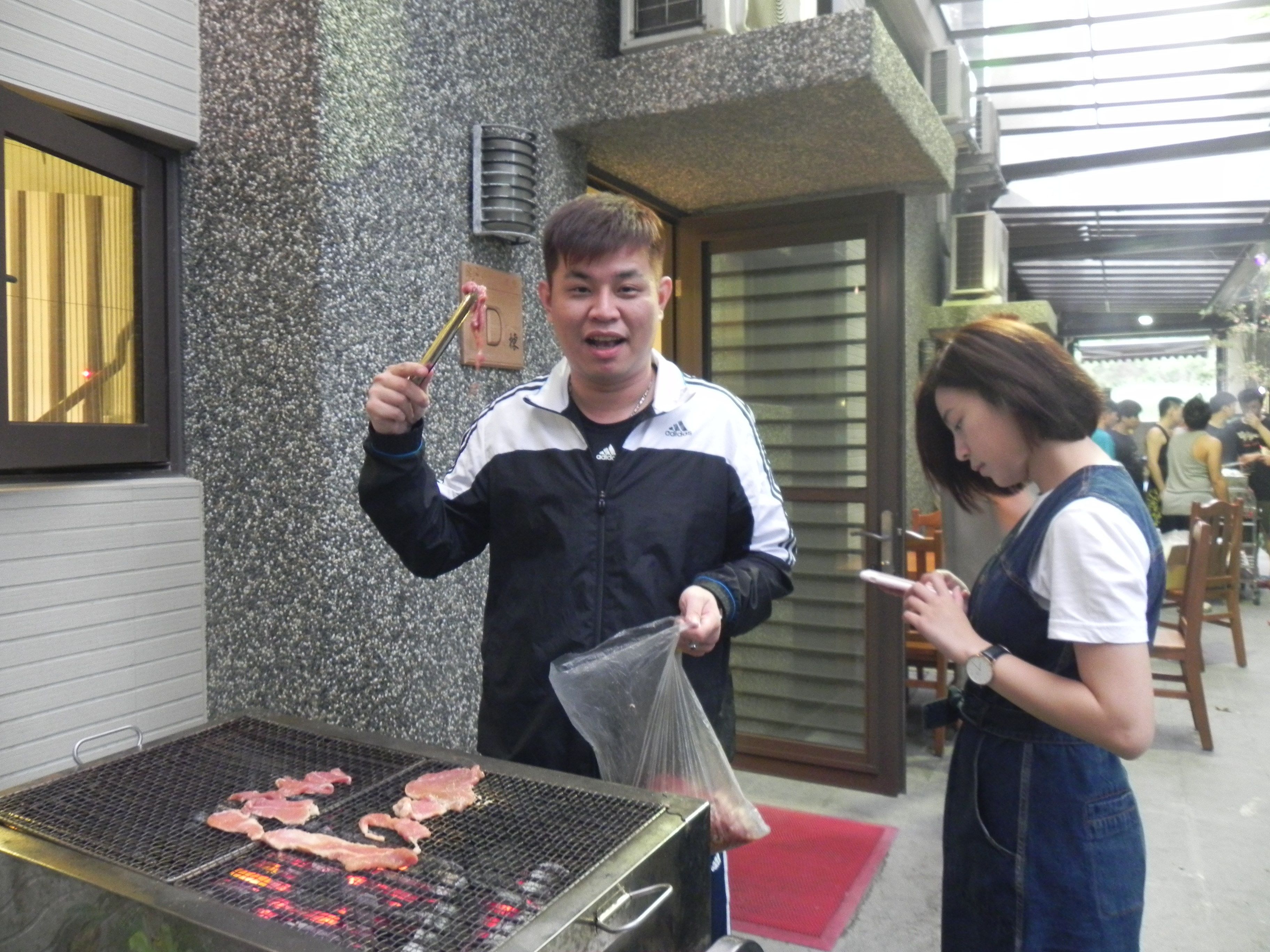 A person grilling meat on an outdoor grill with a stone wall and a window in the background.