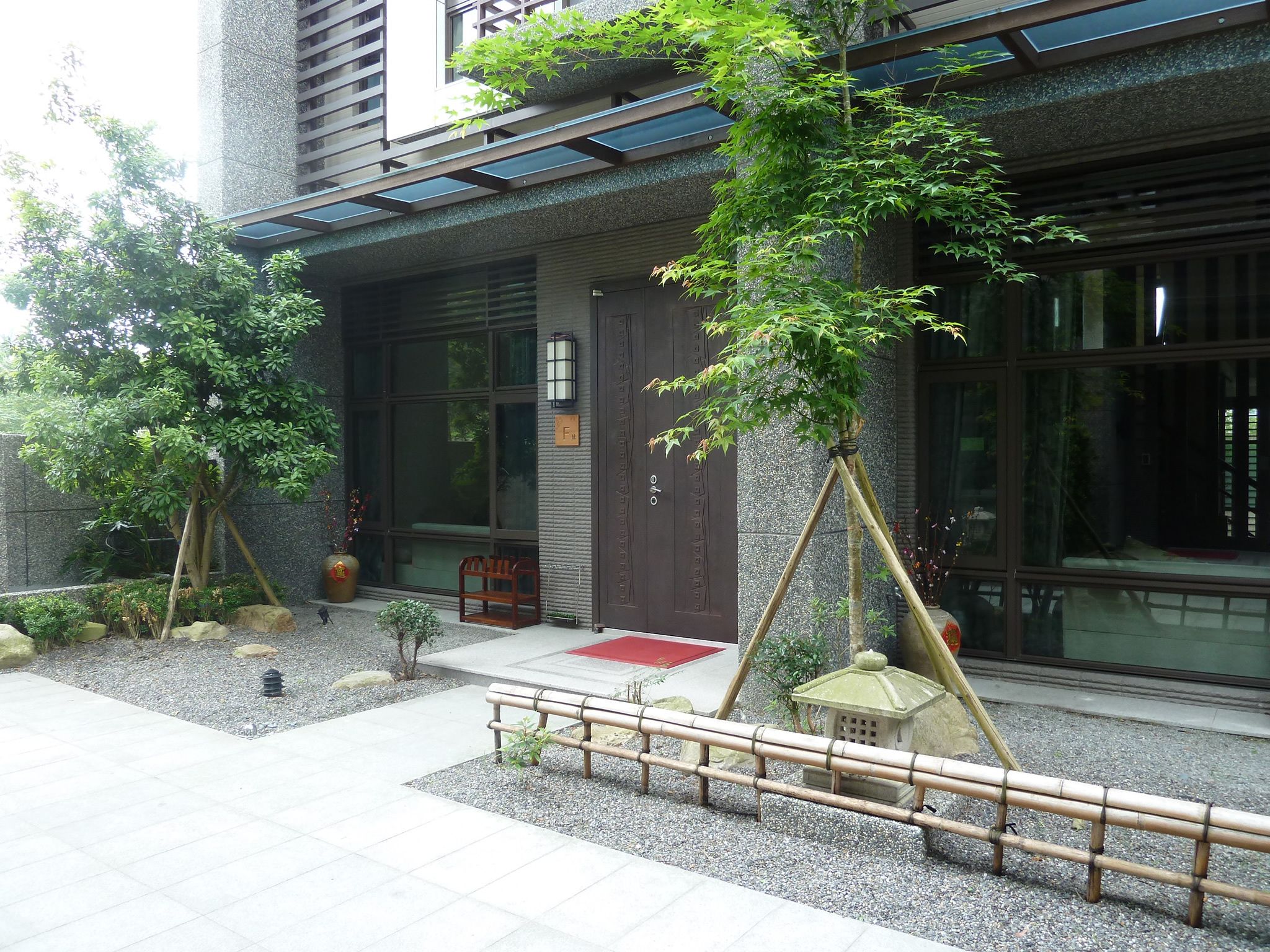 A traditional Japanese building with a wooden door, a red mat, and a small garden area with a bamboo fence and a stone lantern.
