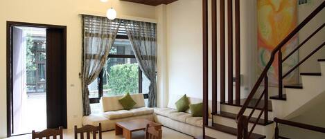 A modern living room with a sofa, dining table, and chairs, a staircase, and a large window with curtains.