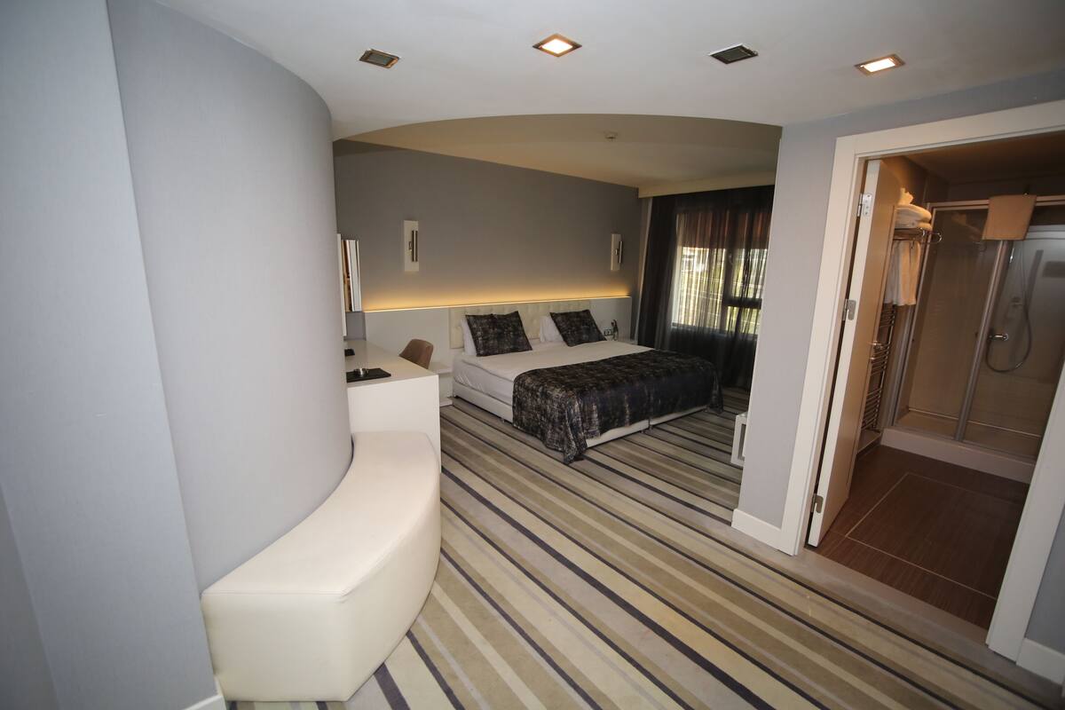 Une chambre d’hôtel moderne avec un banc incurvé, un grand lit surmonté d’un tapis à rayures, une douche à l’italienne et une vue sur l’extérieur.