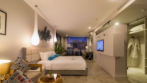 Una habitación de hotel moderna con una cama grande, mesita de noche, lámpara y televisor. Cuenta con una zona de estar con una mesa y sillas, y una bata colgada en la pared.