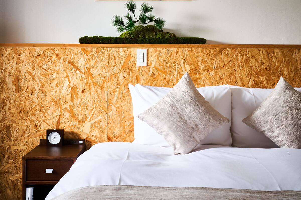 Habitación de hotel con cabecera de madera, cama con ropa blanca, mesita de noche con un reloj y una obra de arte colgada en la pared.