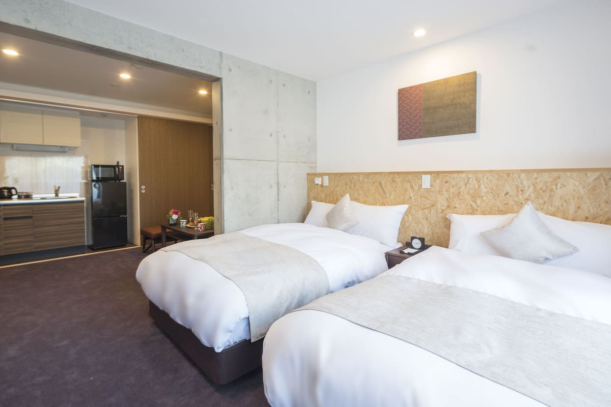 Habitación de hotel con dos camas, cabecera de madera, un cuadro en la pared y una kitchenette al fondo.