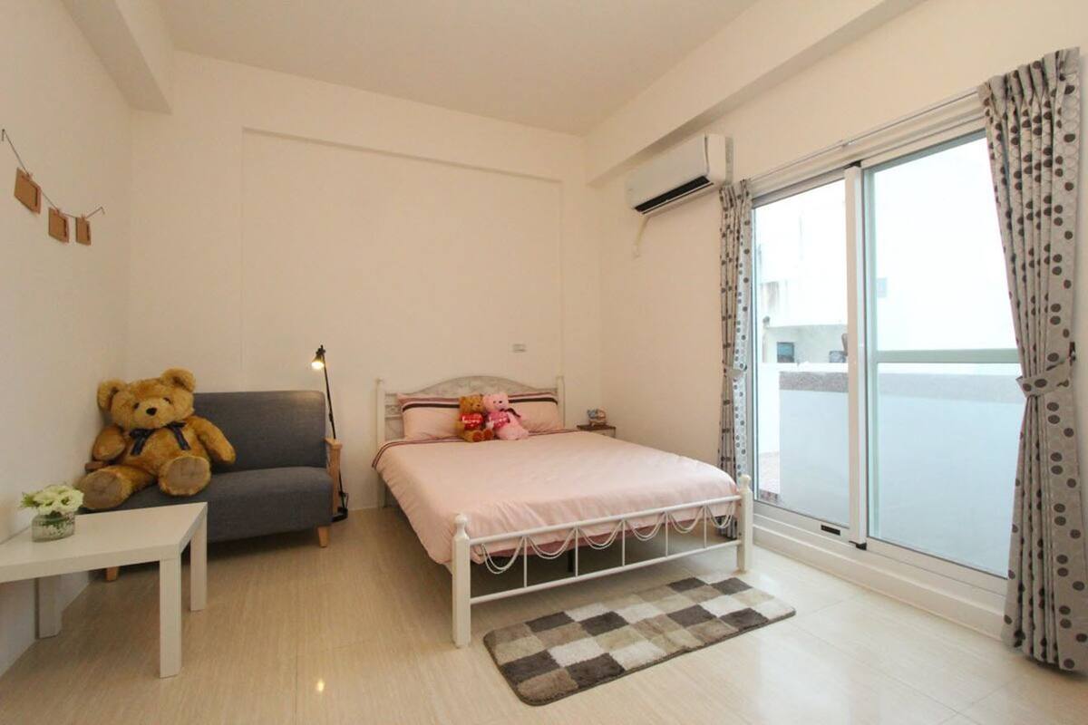 A bedroom with a bed, a sofa, a teddy bear, a small table, and a window with curtains.