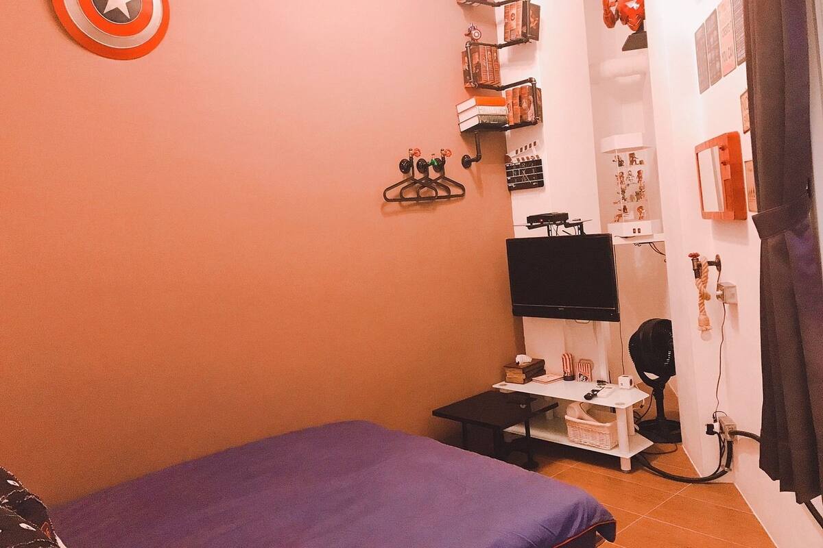 A bedroom with a bed, a desk with a computer, a TV, and a shelf with books and decorations.