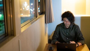 A person sitting at a table with a laptop, near a window with curtains, in a room with a lamp and a view of city lights outside. - Hotel S-Presso North (Osaka)