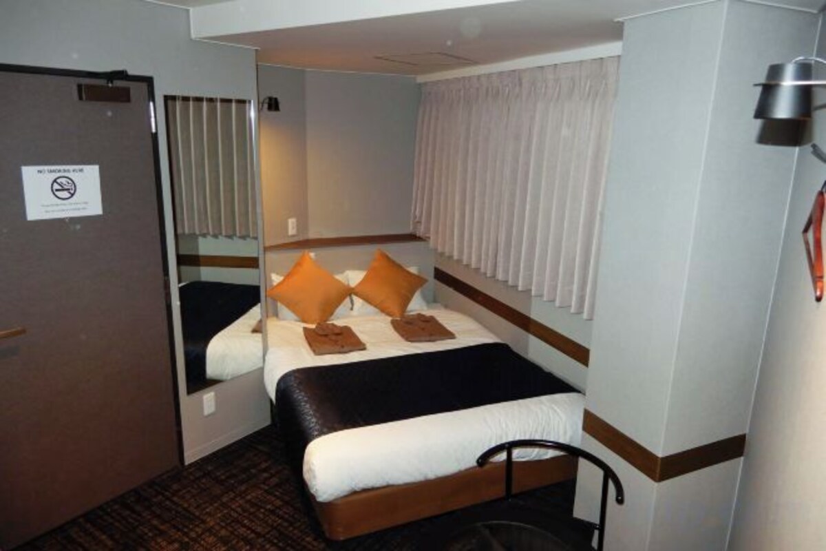 A hotel room with a bed, a door, a window with curtains, and a wall-mounted light fixture.
