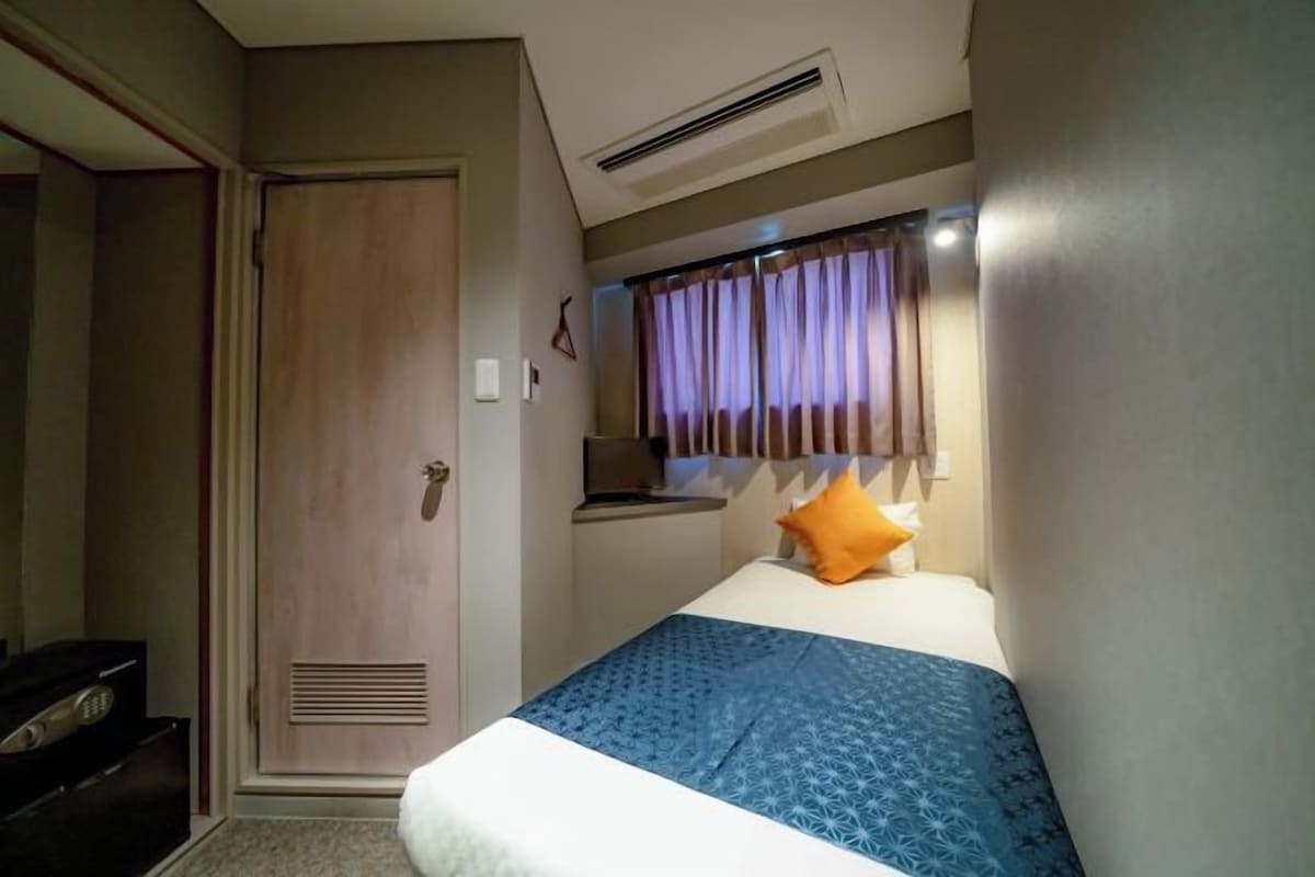 A hotel room with a bed, a bedside table, a window with curtains, a door, and a built-in air conditioning unit.