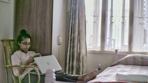 A person sitting on a wicker chair with an HP laptop, in a room with a bed, curtains, and a window with vertical blinds. - Casa Mia (Guwahati)