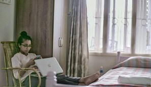 A person sitting on a wicker chair with an HP laptop, in a room with a bed, curtains, and a window with vertical blinds. - Casa Mia (Guwahati)