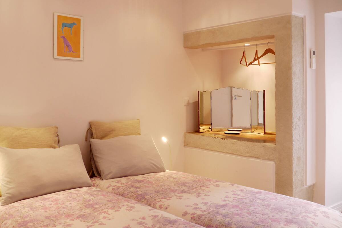 A bedroom with a bed, two pillows, a bedside table, a nightstand, a mirror, and a wall-mounted artwork.