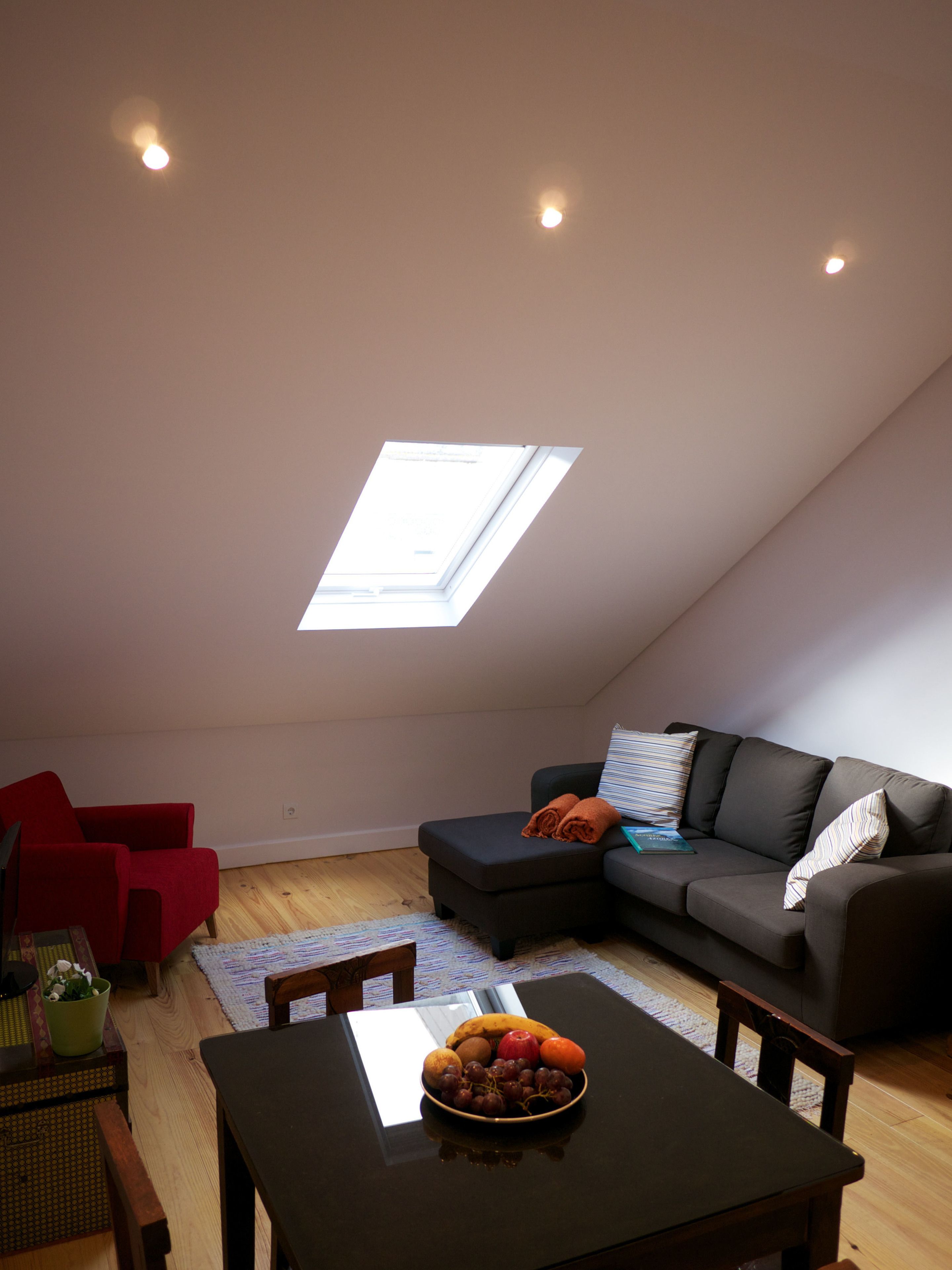 A modern loft apartment with a skylight, a dining area with a fruit bowl, a red armchair, and a dark grey sofa.