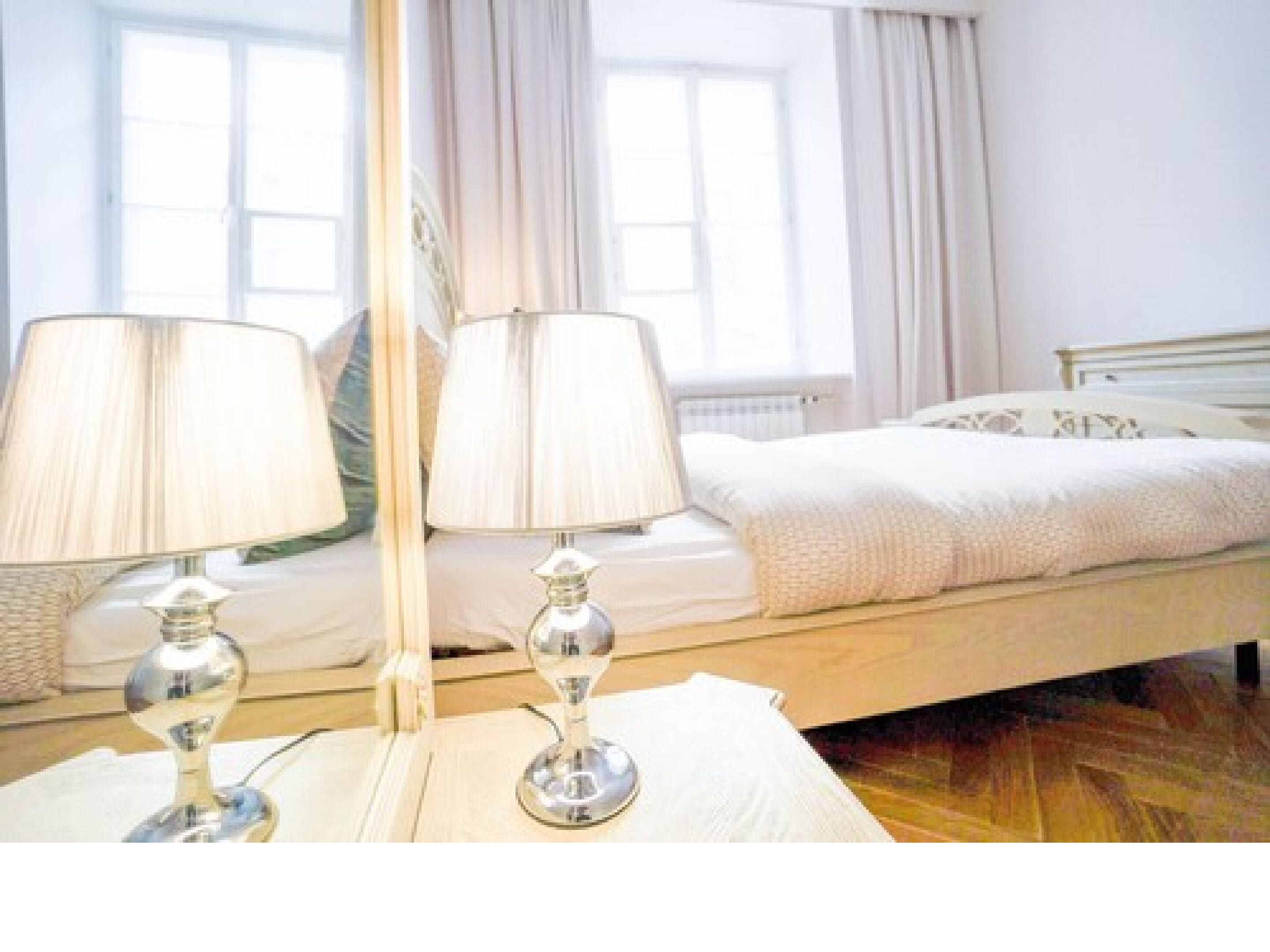 A neatly made bed with white linens and a wooden headboard, two bedside lamps, and a window with curtains.