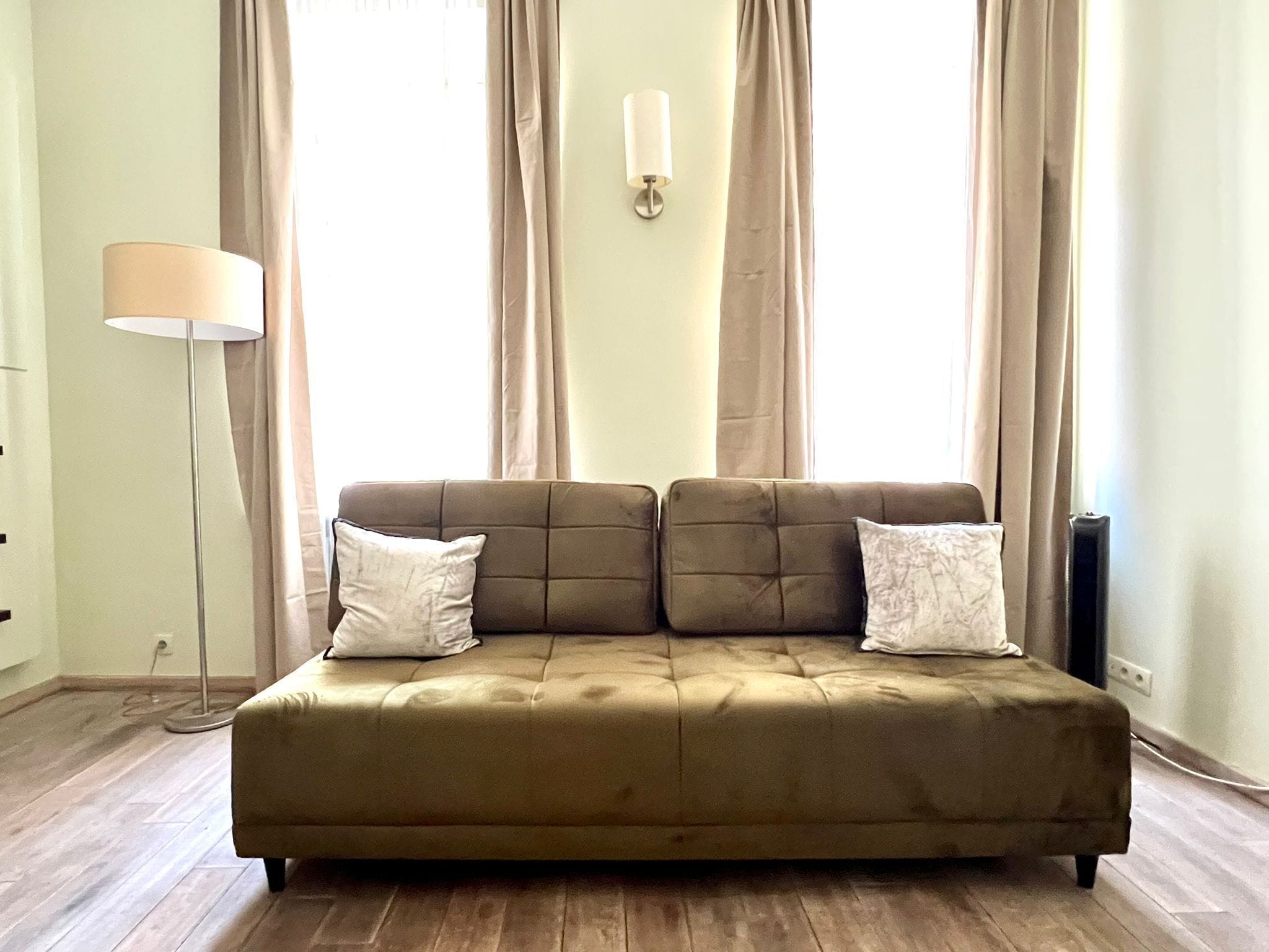 Um sofá bege com duas almofadas, um abajur e um luminário fixado na parede, em um quarto com janelas amplas e cortinas.