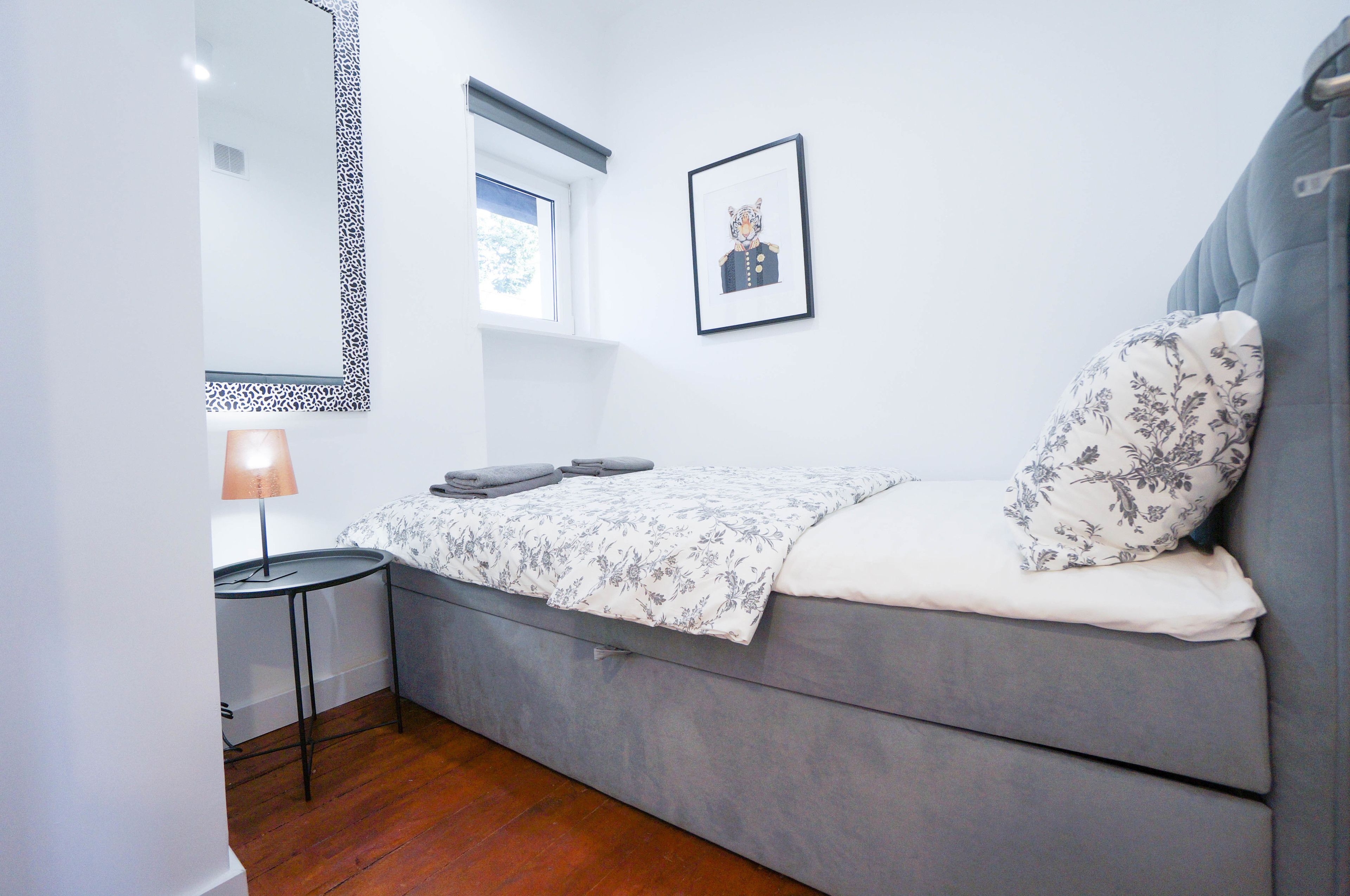 Une petite chambre à coucher, bien rangée, avec un canapé-lit gris, une petite table avec une lampe, un tableau encadré au mur et une fenêtre donnant sur des arbres.