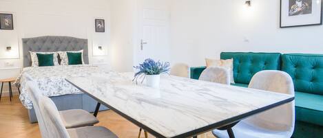 A modern living room with a large sofa, a dining table with chairs, a bed with a tufted headboard, and framed artwork on the walls.
