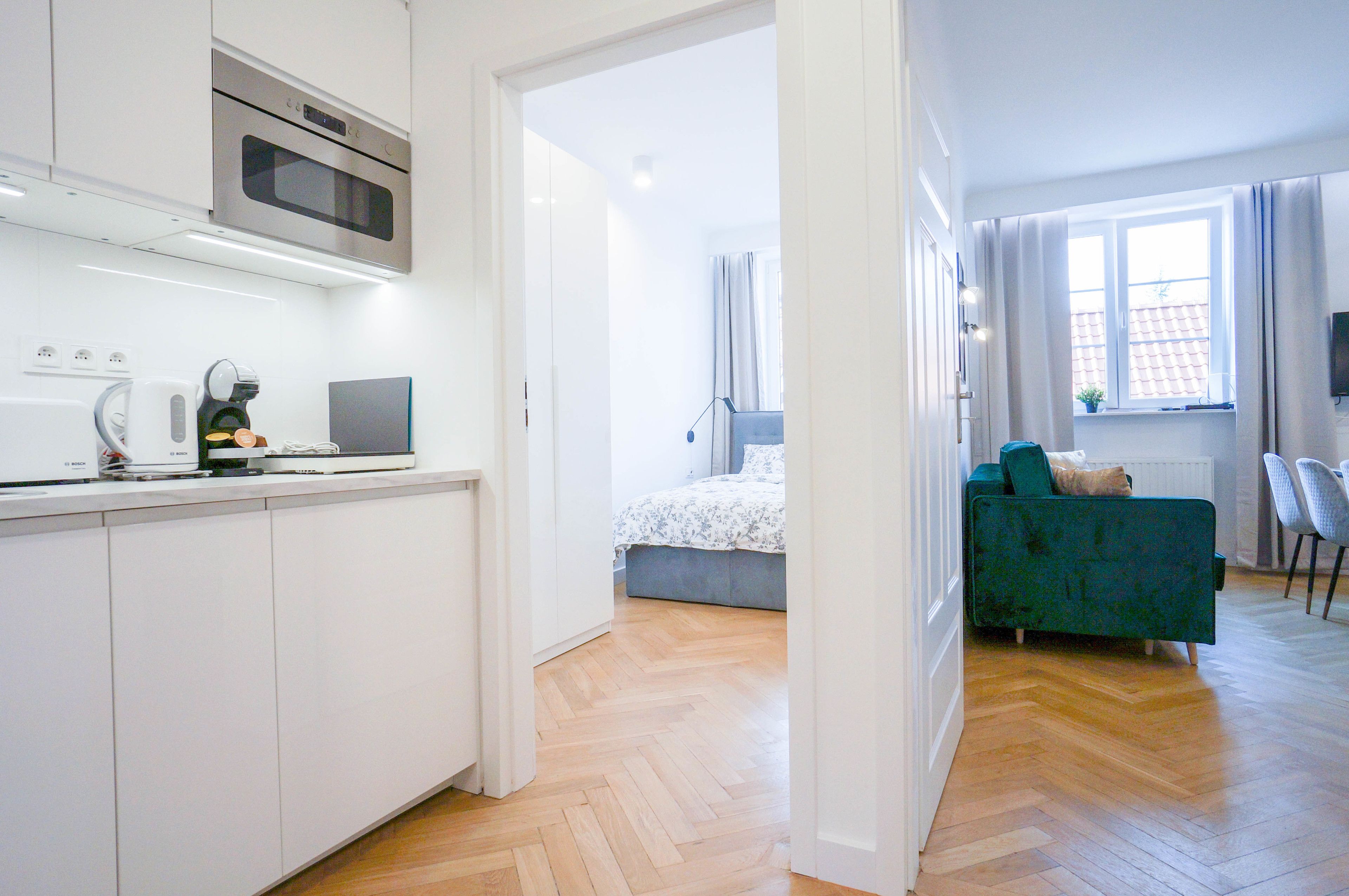Una cucina moderna con mobili bianchi e forno integrato, una camera da letto con un letto e una finestra, e un soggiorno con un divano verde e un tavolino da caffè.