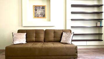 Un sofá marrón con dos almohadas, un cuadro con la palabra “Love” en la pared y un estante de madera oscura adosado a la pared.